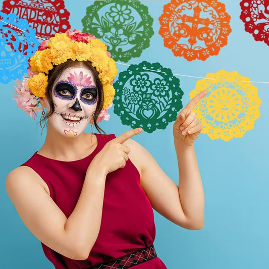 A woman with Day of the Dead sugar skull face paint wears a yellow flower crown and points to colorful papel picado banners hanging on a light blue background.