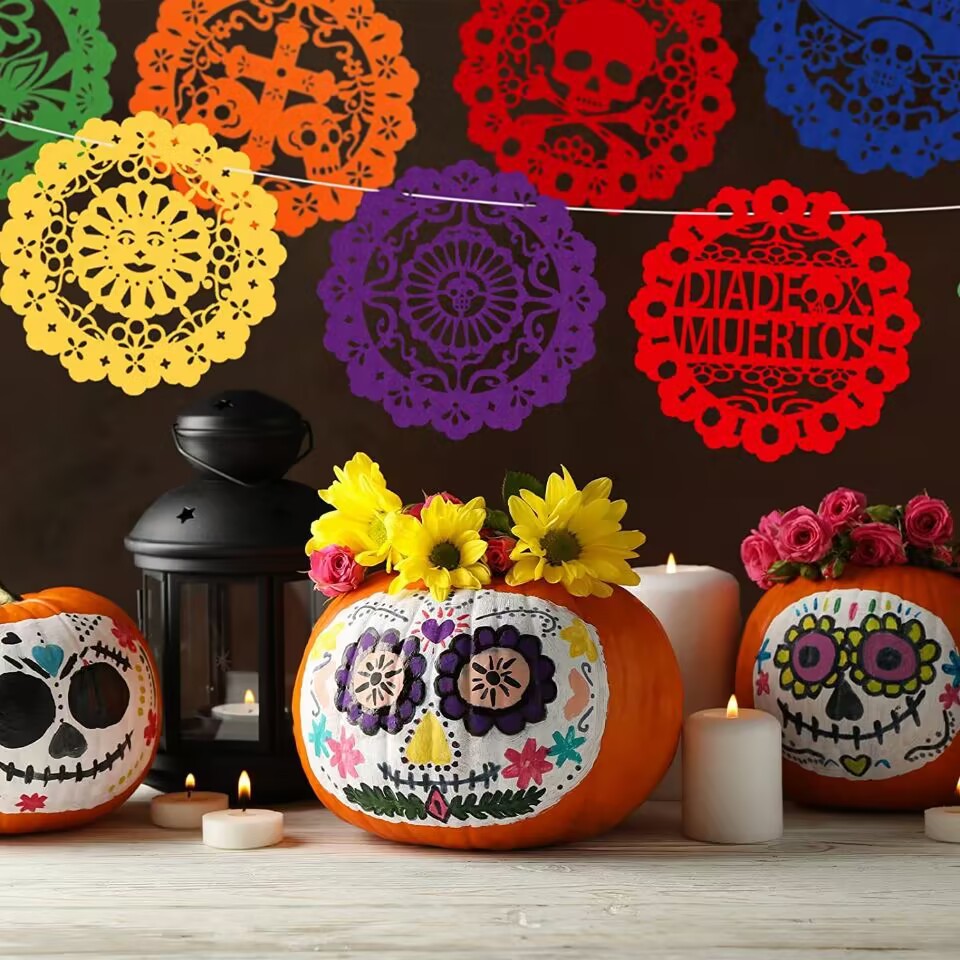 Three decorated orange pumpkins with colorful sugar skull designs and flower arrangements, displayed on a wooden surface with candles and a black lantern. Colorful papel picado banners hang on a dark brown wall behind them.