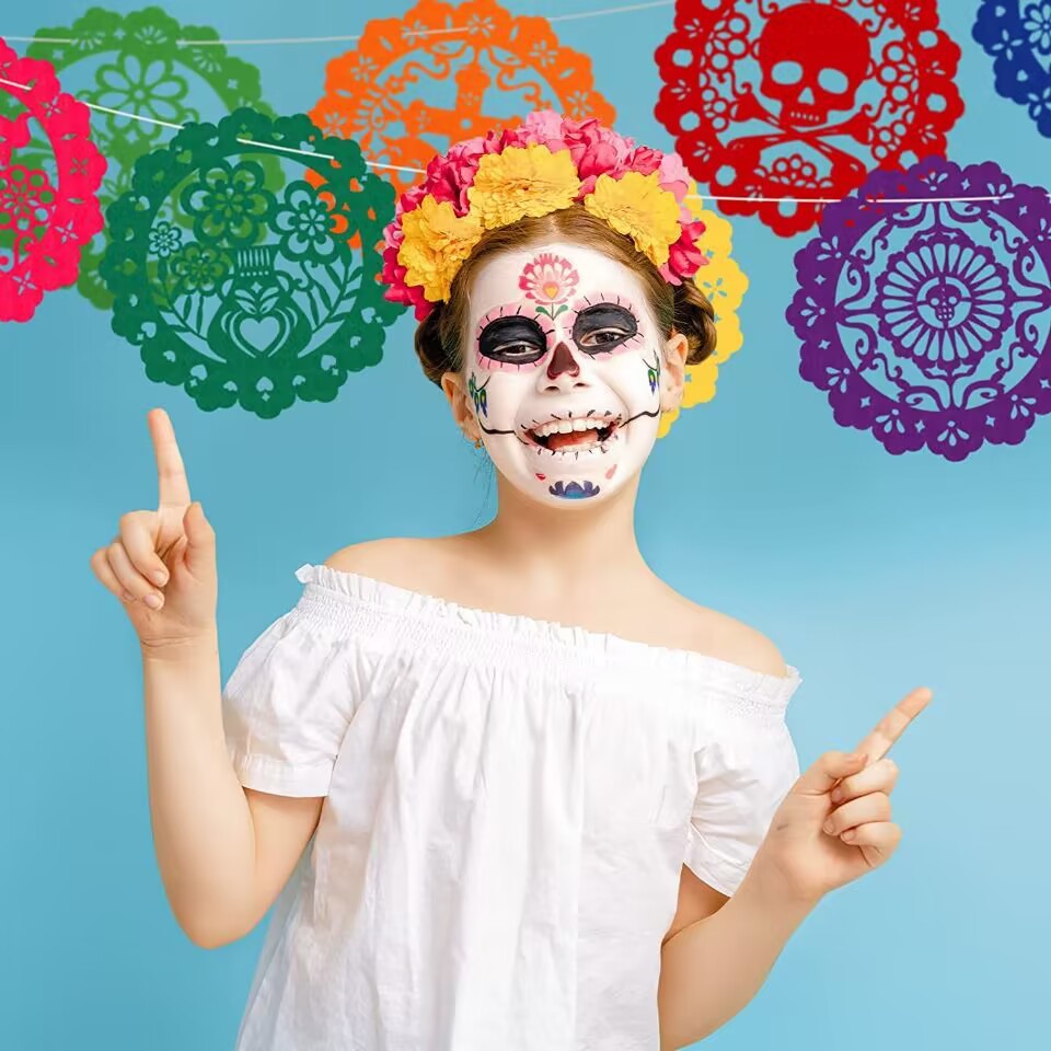 A young girl dressed in Day of the Dead (Dia de los Muertos) attire, featuring sugar skull face makeup, a yellow and pink floral crown, and a white off-the-shoulder top, pointing upwards against a light blue background with colorful papel picado banners.