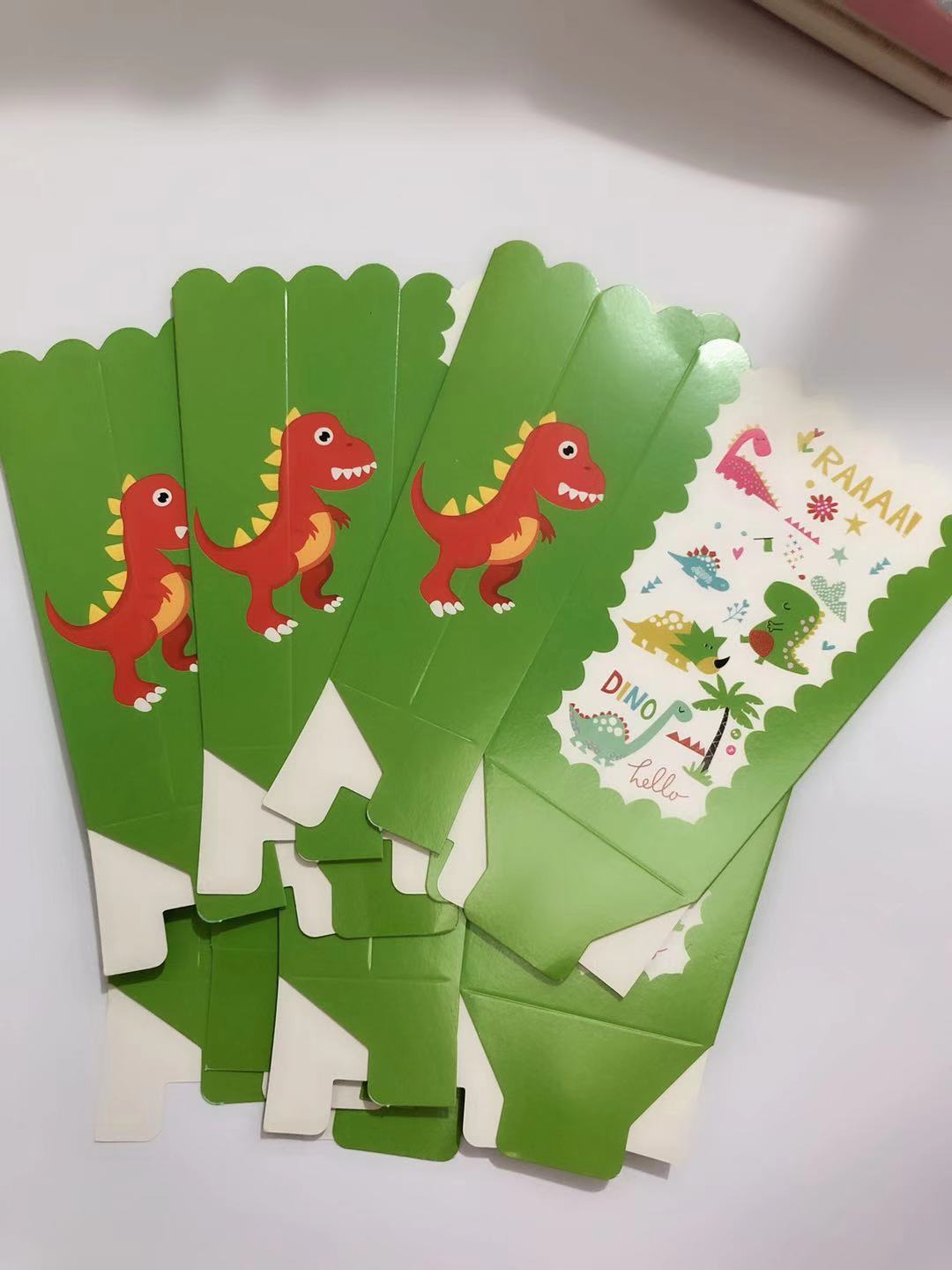 A set of green dinosaur-themed party cupcake toppers or packaging pieces fanned out on a white surface. The pieces feature cartoon red and yellow dinosaurs, palm trees