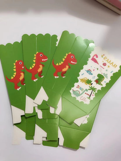 A set of green dinosaur-themed party cupcake toppers or packaging pieces fanned out on a white surface. The pieces feature cartoon red and yellow dinosaurs, palm trees