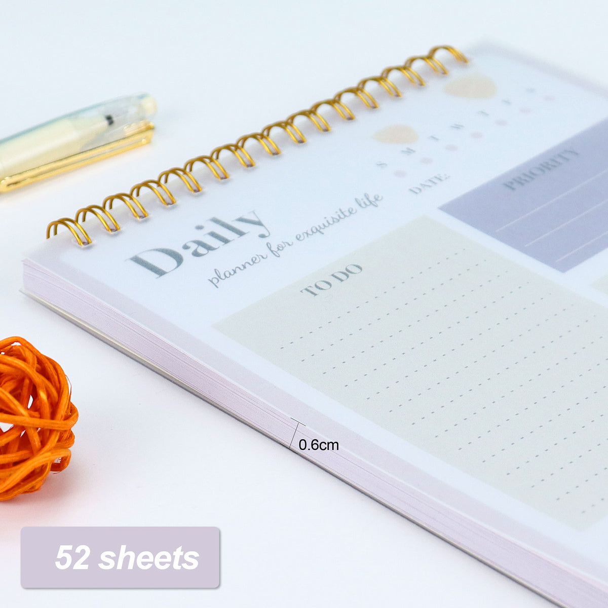 A spiral-bound daily planner notebook shown open on a white surface, with a gold wire spiral binding, pastel planner pages, and a small printed overlay indicating 52 sheets and 0.6cm sheet thickness.