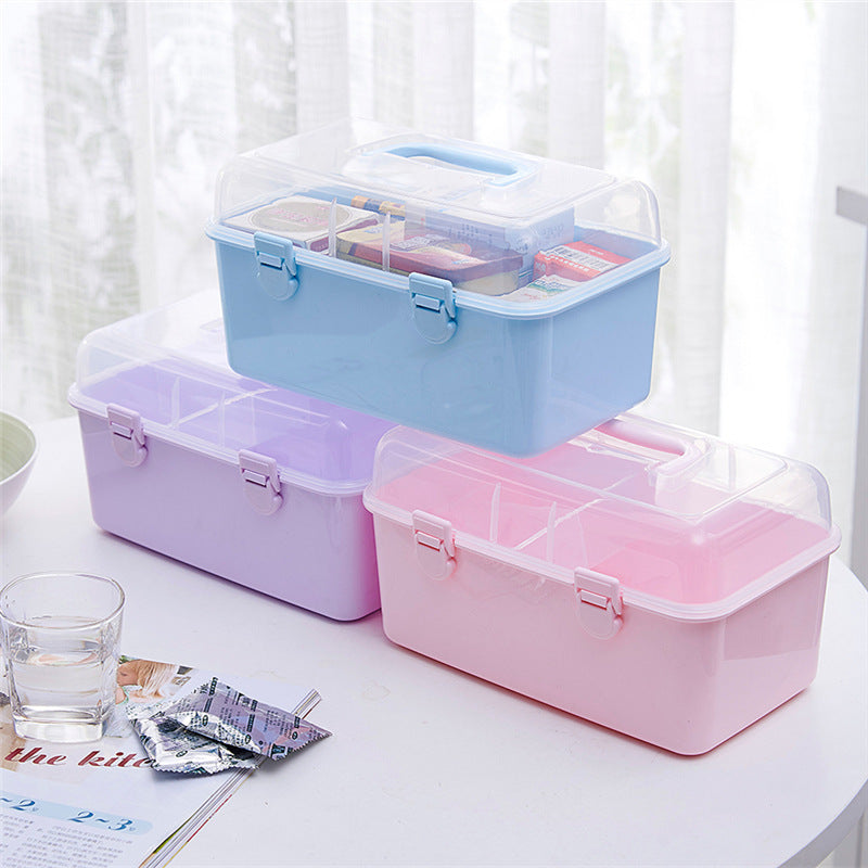 Three plastic storage boxes stacked on a white table, with a blue box on top containing small compartments and items, a lavender box in the middle, and a pink box on the bottom. The boxes have clear lids and are shown in a bright indoor setting.