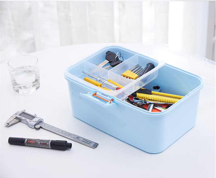A light blue plastic organizer box with a clear divided lid, filled with various tools, placed on a white table next to a glass of water, a caliper, and a marker.